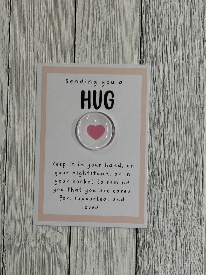 Puede incluir: Una tarjeta blanca con borde rosa dice "Sending you a HUG". Un peque&ntilde;o c&iacute;rculo transparente con un coraz&oacute;n rosa en su interior est&aacute; unido a la tarjeta. El texto debajo del coraz&oacute;n dice "Keep it in your hand, on your nightstand, or in your pocket to remind you that you are cared for, supported, and loved."