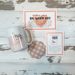 May include: A white ceramic mug with the words "hug in a rug" and a watercolor design, a coaster, and two cards with text. The cards read "Sending you a Big Warm Hug" and "Sending you a Hug". The coaster has a pink and white checkered pattern.