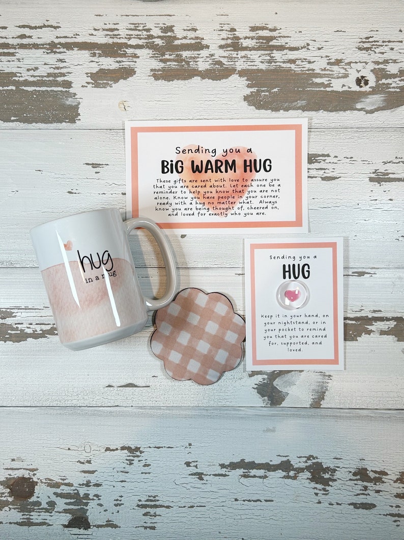 May include: A white ceramic mug with the words "hug in a rug" and a watercolor design, a coaster, and two cards with text. The cards read "Sending you a Big Warm Hug" and "Sending you a Hug". The coaster has a pink and white checkered pattern.