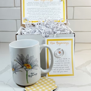 Pu&ograve; includere: Una tazza bianca con un disegno di margherita gialla e bianca e il testo "New Beginnings". La tazza &egrave; appoggiata su un sottobicchiere a scacchi giallo e bianco. C'&egrave; un piccolo biglietto con il testo "New Beginnings" e un disegno di margherita.