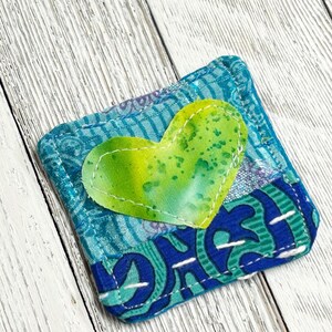 May include: A small, blue and green fabric pouch with a heart-shaped patch sewn on the front. The heart is green with yellow and white speckles.