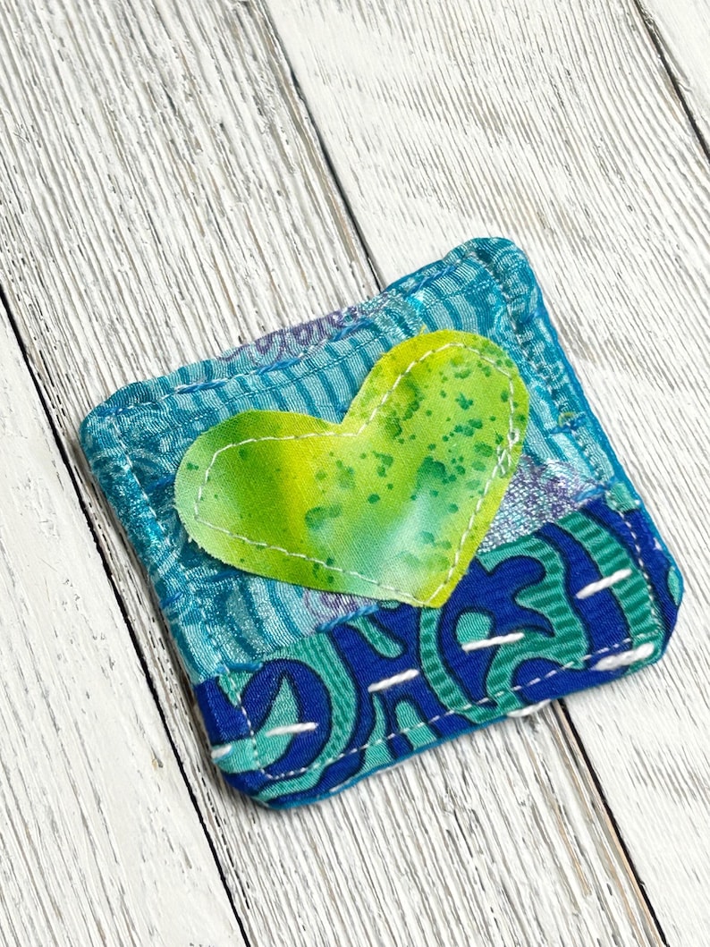 May include: A small, blue and green fabric pouch with a heart-shaped patch sewn on the front. The heart is green with yellow and white speckles.