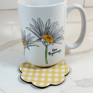 Pu&ograve; includere: Tazza in ceramica bianca con un design di margherita ad acquerello e il testo "Nuovi Inizi". La tazza &egrave; appoggiata su un sottobicchiere a quadri gialli e bianchi.