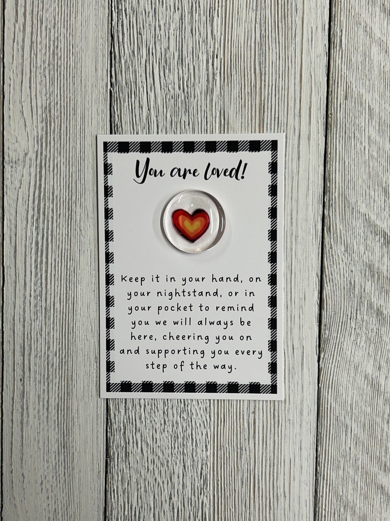 K&ouml;nnte beinhalten: Eine wei&szlig;e Karte mit einem schwarz-wei&szlig; karierten Rand und den Worten "You are loved!". Ein Glashherz mit roten und gelben Farbt&ouml;nen ist unter dem Text zentriert. Zus&auml;tzlicher Text ermutigt den Empf&auml;nger, es als Erinnerung an die Unterst&uuml;tzung zu behalten.