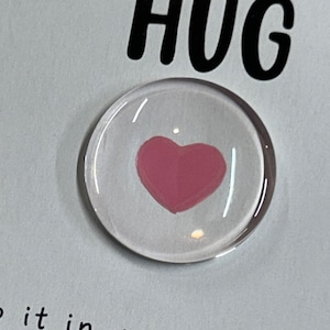 Puede incluir: Un peque&ntilde;o c&iacute;rculo de vidrio con un coraz&oacute;n rosa en su interior. El texto "Sending you a HUG" es visible en el fondo.