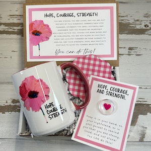 May include: A gift box filled with a white mug with a pink poppy design and the words "Hope Courage Strength" on it. The box also includes a pink and white checkered coaster, a pink heart-shaped stone, and a card with the words "Hope Courage and Strength" and the text "Keep it in your hand, on your nightstand, or in your pocket to remind you that you are strong, loved, and not alone! You CAN do this!"