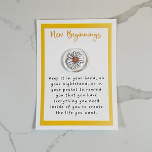 Pu&ograve; includere: Un biglietto giallo con il testo "New Beginnings" e una margherita bianca su uno sfondo trasparente. Il testo sotto la margherita dice "Keep it in your hand, on your nightstand, or in your pocket to remind you that you have everything you need inside of you to create the life you want."