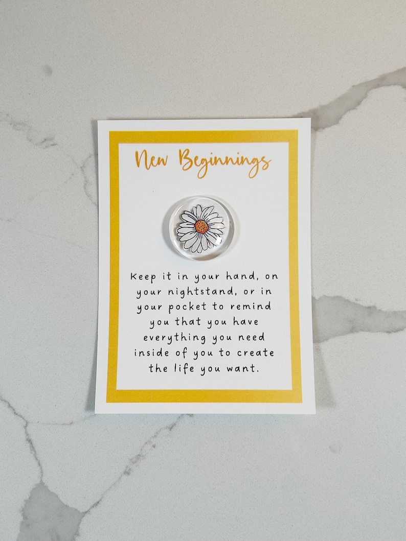 Pu&ograve; includere: Un biglietto giallo con il testo "New Beginnings" e una margherita bianca su uno sfondo trasparente. Il testo sotto la margherita dice "Keep it in your hand, on your nightstand, or in your pocket to remind you that you have everything you need inside of you to create the life you want."