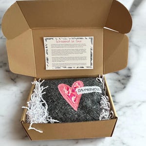 May include: A brown cardboard box with a gray fabric heart-shaped pillow inside. The pillow has a pink heart with white polka dots sewn on it. The pillow has the word "strength" written on it. The box is filled with white shredded paper.