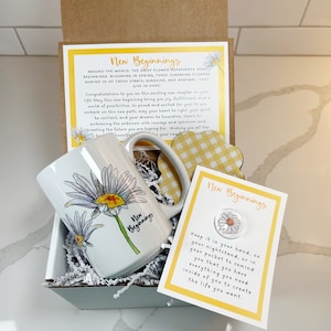 Pu&ograve; includere: Un set regalo con una tazza bianca con un motivo di margherita e il testo "Nuovi Inizi". La scatola include anche un piccolo biglietto con il testo "Nuovi Inizi" e un ciondolo a forma di margherita. La scatola &egrave; riempita con carta triturata bianca e carta a quadri gialla e bianca.