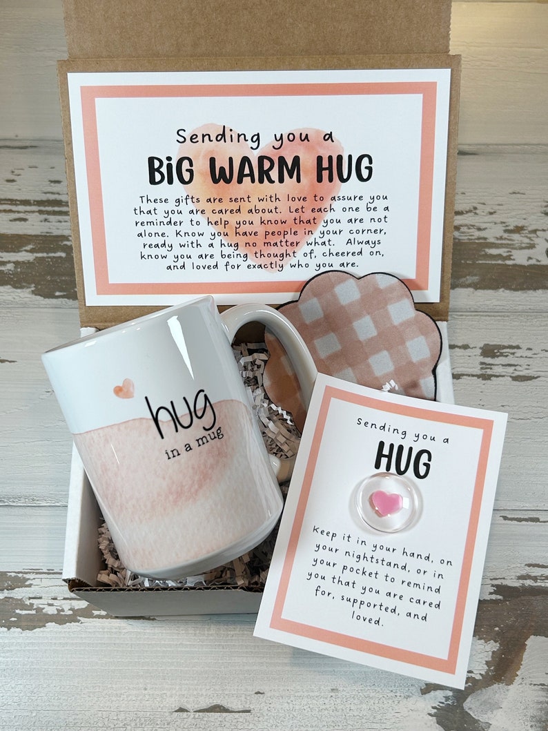May include: A gift box containing a white mug with "hug in a mug" text and a pink watercolor design, a heart-shaped pin, and a decorative cloud. The box includes a card with the text "Sending you a Big Warm Hug."
