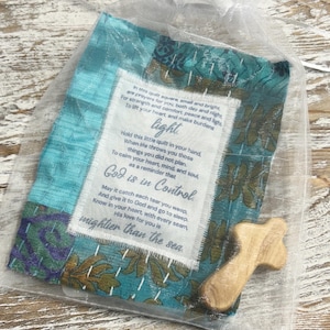 May include: A small teal blue fabric square with a white tag attached. The tag has a poem about faith and hope. The fabric square is in a clear organza bag with a small wooden cross. The poem reads: "In this quilt square, small and bright are prayers for you both day and night For strength and comfort, peace and light, To lift your heart, and make burdens light Hold this little quilt in your hand, When life throws you those things you did not plan. To calm your heart, mind, and soul, as a reminder that God is in Control May it catch each tear you weep, And give it to God and go to sleep. Know in your heart, with every seam, His love for you is mightier than the sea."