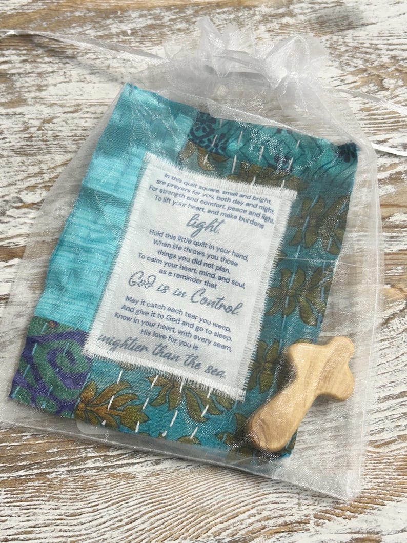 May include: A small teal blue fabric square with a white tag attached. The tag has a poem about faith and hope. The fabric square is in a clear organza bag with a small wooden cross. The poem reads: "In this quilt square, small and bright are prayers for you both day and night For strength and comfort, peace and light, To lift your heart, and make burdens light Hold this little quilt in your hand, When life throws you those things you did not plan. To calm your heart, mind, and soul, as a reminder that God is in Control May it catch each tear you weep, And give it to God and go to sleep. Know in your heart, with every seam, His love for you is mightier than the sea."