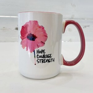 May include: White ceramic mug with a pink flower graphic and the words "Hope Courage Strength" printed in black.