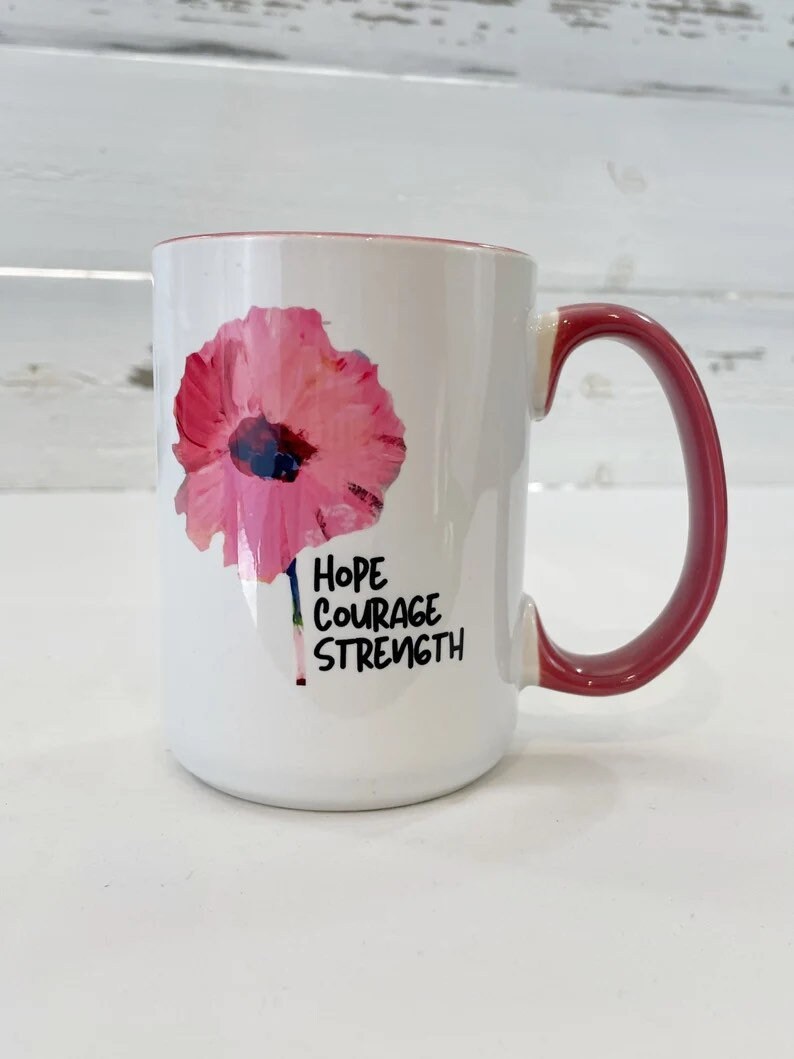 May include: White ceramic mug with a pink flower graphic and the words "Hope Courage Strength" printed in black.