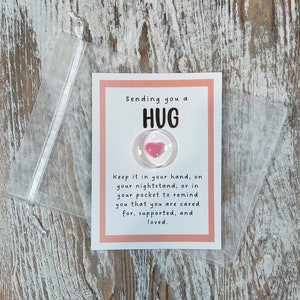May include: A small card with the words "Sending you a HUG" and a clear, round charm with a pink heart. The card is in a clear, plastic pouch. The text on the card reads, "Keep it in your hand, on your nightstand, or in your pocket to remind you that you are cared for, supported, and loved."
