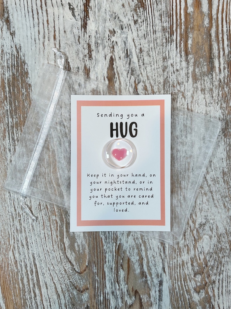 May include: A small card with the words "Sending you a HUG" and a clear, round charm with a pink heart. The card is in a clear, plastic pouch. The text on the card reads, "Keep it in your hand, on your nightstand, or in your pocket to remind you that you are cared for, supported, and loved."