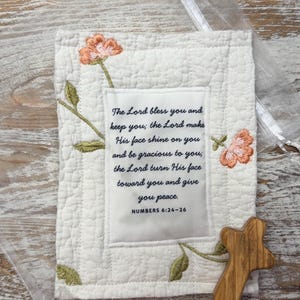 May include: A rectangular white quilted fabric panel featuring embroidered peach flowers and green stems. A central white fabric patch displays the black text: The Lord bless you and keep you; the Lord make His face shine on you and be gracious to you; the Lord turn His face toward you and give you peace. NUMBERS 6:24-26. A small wooden cross is placed at the bottom right.