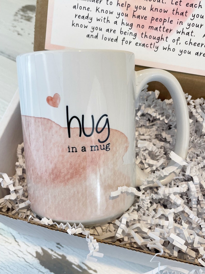 May include: White ceramic mug with a watercolor-style design. The mug features a pink heart and the words "hug in a mug" in black script. The mug is presented in a cardboard box with white paper filler, suggesting a gift.