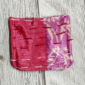 May include: A small, square, pink and white fabric pouch with white stitching around the edges. The pouch is made of two different fabrics, one with a floral pattern and the other with a solid pink color.