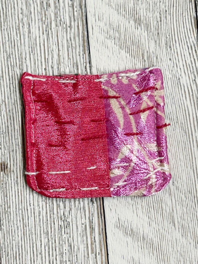 May include: A small, square, pink and white fabric pouch with white stitching around the edges. The pouch is made of two different fabrics, one with a floral pattern and the other with a solid pink color.