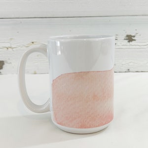 May include: White ceramic mug with a large handle. The bottom half of the mug features a watercolor-style design in shades of peach and pink. The mug is set against a white background.