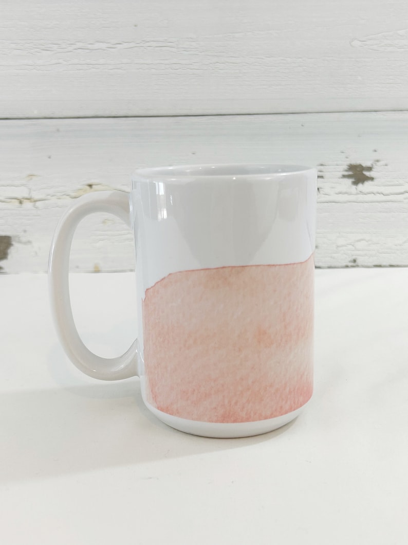 May include: White ceramic mug with a large handle. The bottom half of the mug features a watercolor-style design in shades of peach and pink. The mug is set against a white background.