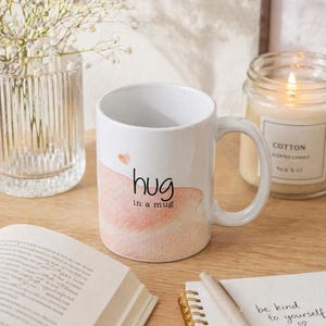 May include: White ceramic mug with the words "hug in a mug" in black script, over a watercolor-style peach-colored design. A small pink heart is above the word "hug". The mug is on a wooden surface.