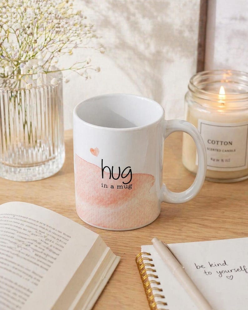 May include: White ceramic mug with the words "hug in a mug" in black script, over a watercolor-style peach-colored design. A small pink heart is above the word "hug". The mug is on a wooden surface.