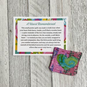 May include: A small pink and green fabric heart-shaped pocket quilt with a message that reads "A Heart Remembered. This small pocket quilt was made to hold close when your heart feels heavy. Inside, you'll find a wooden heart - a quiet reminder of the love that remains, steady and strong, even in absence. On the outside, a soft fabric heart - to remind you that you are held, wrapped in care and compassion. May this little pocket quilt bring you comfort and peace, and may you always feel the warmth of cherished memories and the quiet nearness of love that never truly leaves."