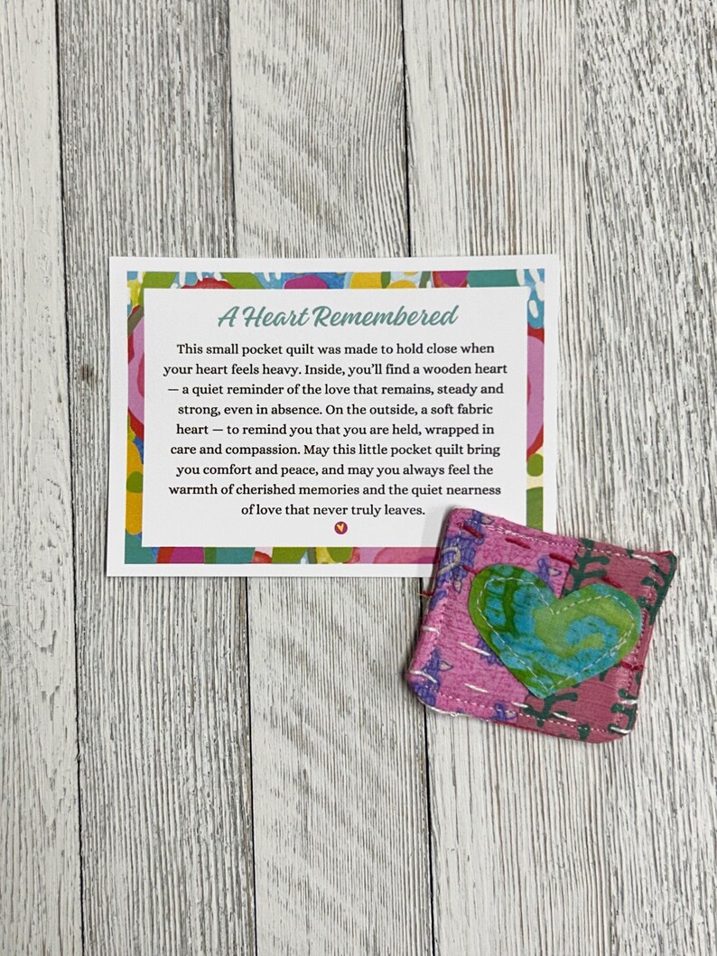 May include: A small pink and green fabric heart-shaped pocket quilt with a message that reads "A Heart Remembered. This small pocket quilt was made to hold close when your heart feels heavy. Inside, you'll find a wooden heart - a quiet reminder of the love that remains, steady and strong, even in absence. On the outside, a soft fabric heart - to remind you that you are held, wrapped in care and compassion. May this little pocket quilt bring you comfort and peace, and may you always feel the warmth of cherished memories and the quiet nearness of love that never truly leaves."
