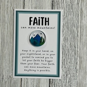 May include: A round, blue, green, and white pin with a mountain and tree design. The pin is on a white card with the words "FAITH can move mountains!" and inspirational text. The card has a teal border.