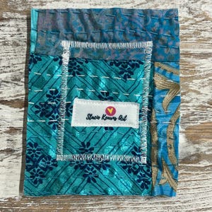 May include: A small, teal blue fabric patchwork with a white label that reads "Stacie Kearns Art" with a pink heart.