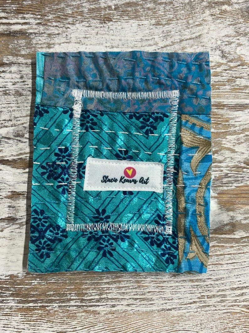 May include: A small, teal blue fabric patchwork with a white label that reads "Stacie Kearns Art" with a pink heart.