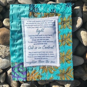 May include: A small, blue and white quilted square with a white fabric label sewn on top. The label has a poem printed on it that reads: "In this quilt square, small and bright, are prayers for you, both day and night. For strength and comfort, peace and light To lift your heart, and make burdens light. Hold this little quilt in your hand, When life throws you those things you did not plan. To calm your heart, mind, and soul as a reminder that God is in Control May it catch each tear you weep, And give it to God and go to sleep. Know in your heart, with every seam, His love for you is mightier than the sea."