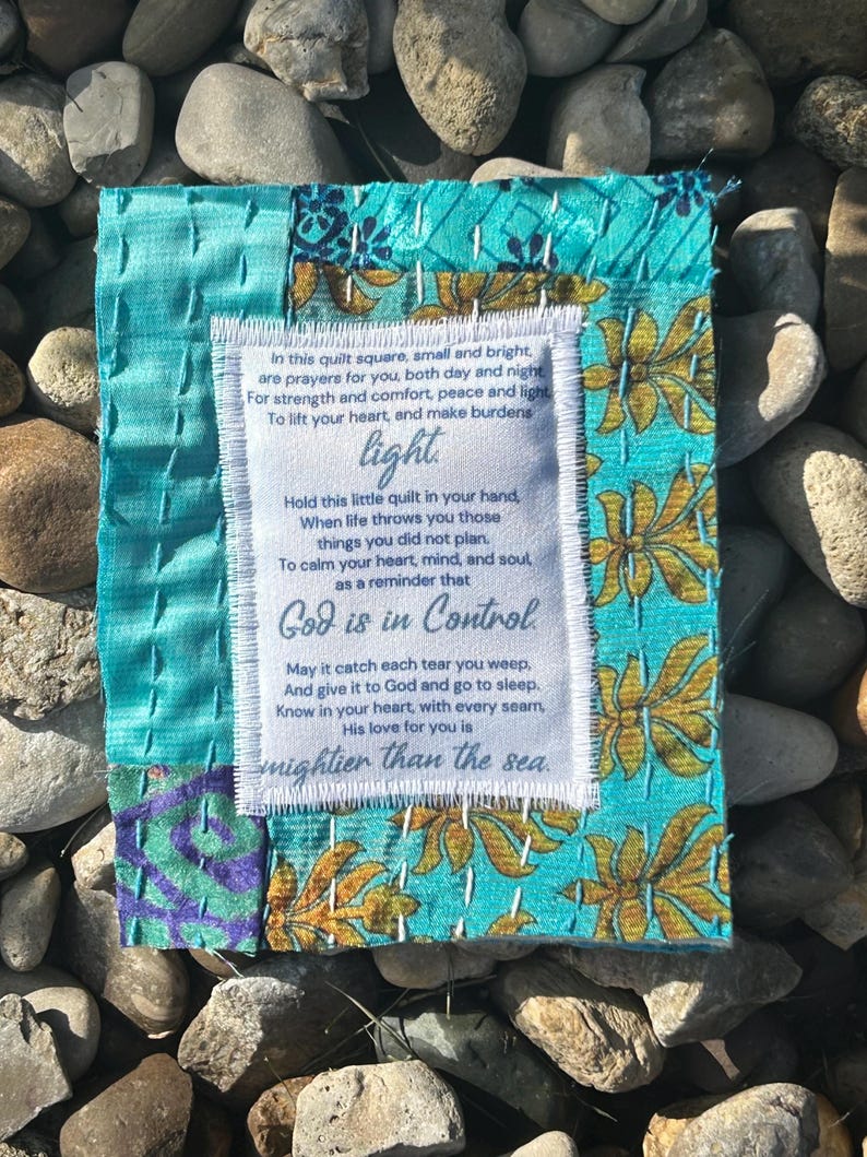 May include: A small, blue and white quilted square with a white fabric label sewn on top. The label has a poem printed on it that reads: "In this quilt square, small and bright, are prayers for you, both day and night. For strength and comfort, peace and light To lift your heart, and make burdens light. Hold this little quilt in your hand, When life throws you those things you did not plan. To calm your heart, mind, and soul as a reminder that God is in Control May it catch each tear you weep, And give it to God and go to sleep. Know in your heart, with every seam, His love for you is mightier than the sea."
