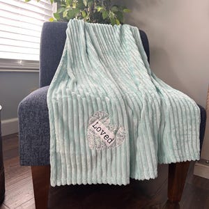 May include: A light blue, ribbed throw blanket draped over a dark blue armchair. The blanket has a heart-shaped patch with the word "Loved" embroidered on it. The chair has wooden legs, and a plant is in the background.