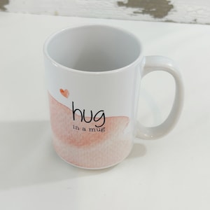 May include: White ceramic mug with a curved handle. The mug features the text "hug in a mug" in black script, with a watercolor-style pink and peach design and a small red heart.