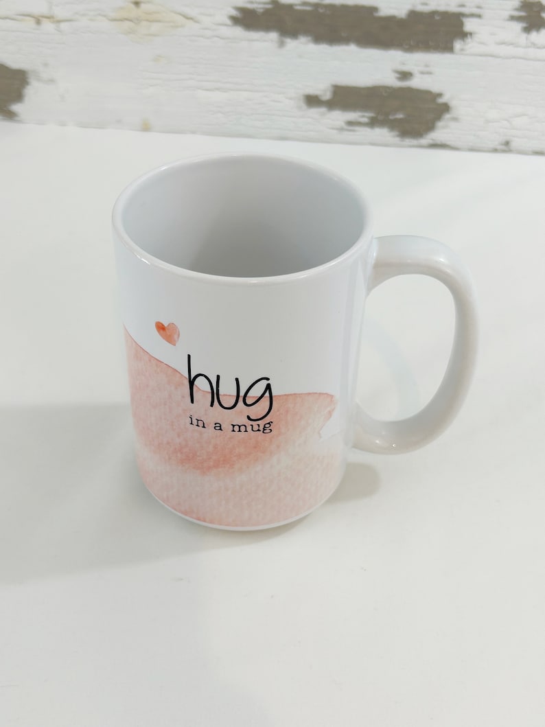 May include: White ceramic mug with a curved handle. The mug features the text "hug in a mug" in black script, with a watercolor-style pink and peach design and a small red heart.