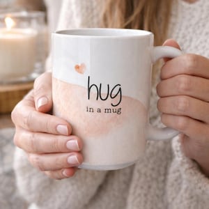 May include: White ceramic mug with a watercolor design in shades of peach and a small heart. The mug has the words "hug in a mug" printed in black. The mug is held by a person.
