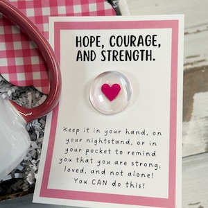 May include: A white card with a pink border and the text "HOPE, COURAGE, AND STRENGTH."  The card also has a small glass heart with a pink heart inside. The text below the heart reads "Keep it in your hand, on your nightstand, or in your pocket to remind you that you are strong, loved, and not alone! You CAN do this!"