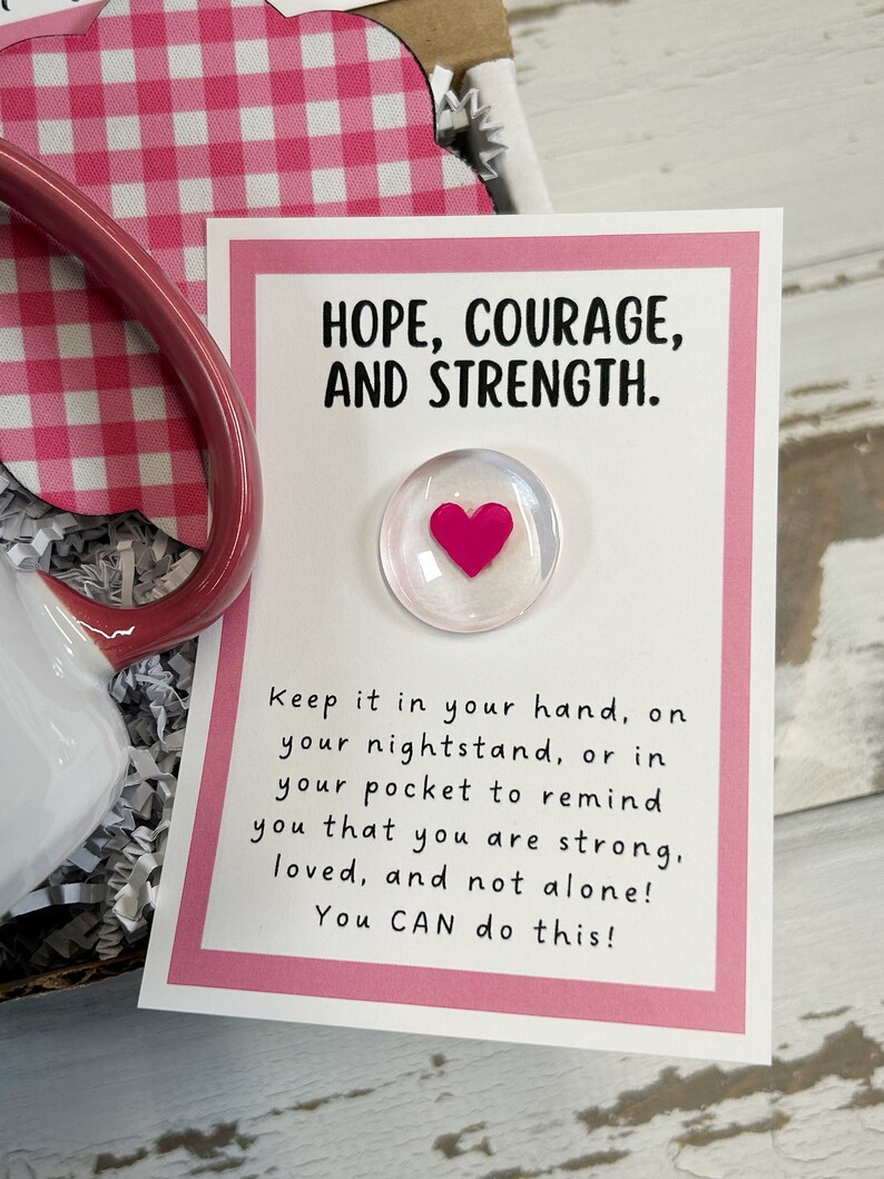 May include: A white card with a pink border and the text "HOPE, COURAGE, AND STRENGTH."  The card also has a small glass heart with a pink heart inside. The text below the heart reads "Keep it in your hand, on your nightstand, or in your pocket to remind you that you are strong, loved, and not alone! You CAN do this!"