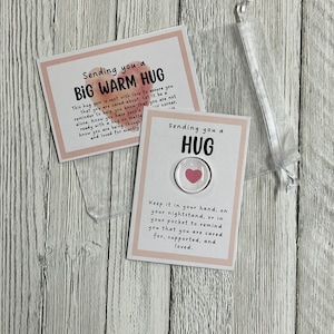 Puede incluir: Dos tarjetas con fondos rosa y blanco. La tarjeta superior dice "Sending you a BIG WARM HUG" y la tarjeta inferior dice "Sending you a HUG". Un peque&ntilde;o amuleto redondo transparente con un coraz&oacute;n rosa en su interior est&aacute; unido a la tarjeta inferior.