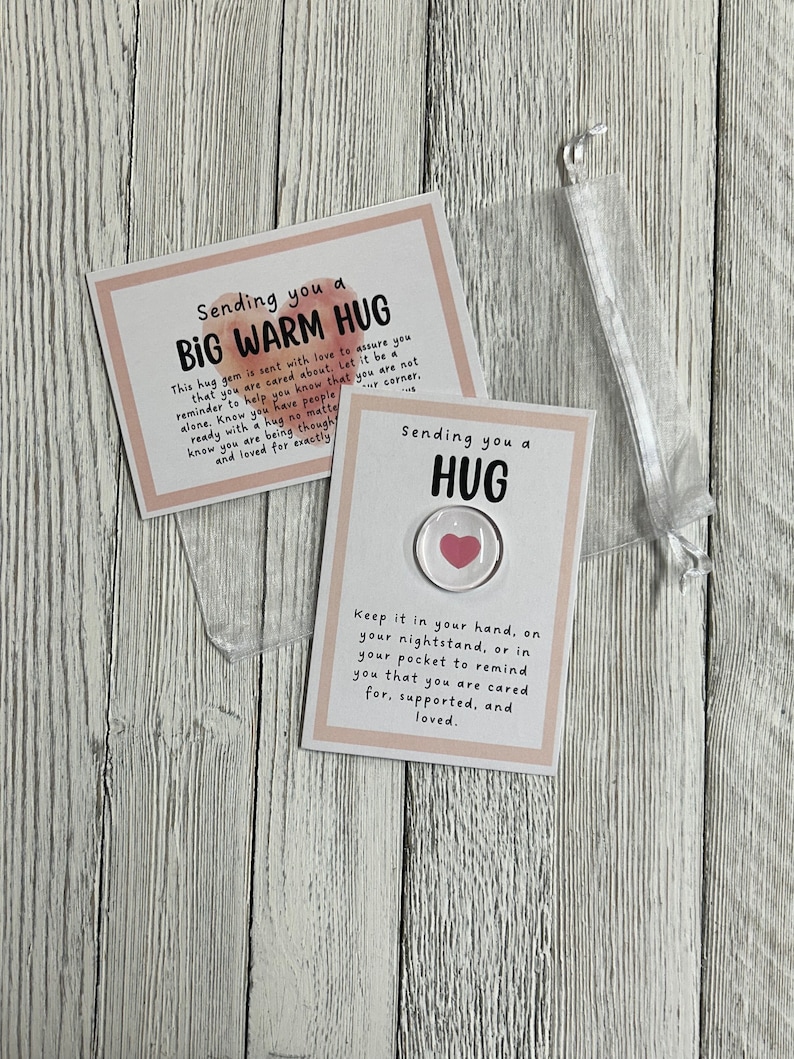 Puede incluir: Dos tarjetas con fondos rosa y blanco. La tarjeta superior dice "Sending you a BIG WARM HUG" y la tarjeta inferior dice "Sending you a HUG". Un peque&ntilde;o amuleto redondo transparente con un coraz&oacute;n rosa en su interior est&aacute; unido a la tarjeta inferior.