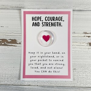 May include: A pink card with the words "HOPE, COURAGE, AND STRENGTH." printed on it. A small glass heart with a pink heart inside is attached to the card. The card reads "Keep it in your hand, on your nightstand, or in your pocket to remind you that you are strong, loved, and not alone! You CAN do this!"