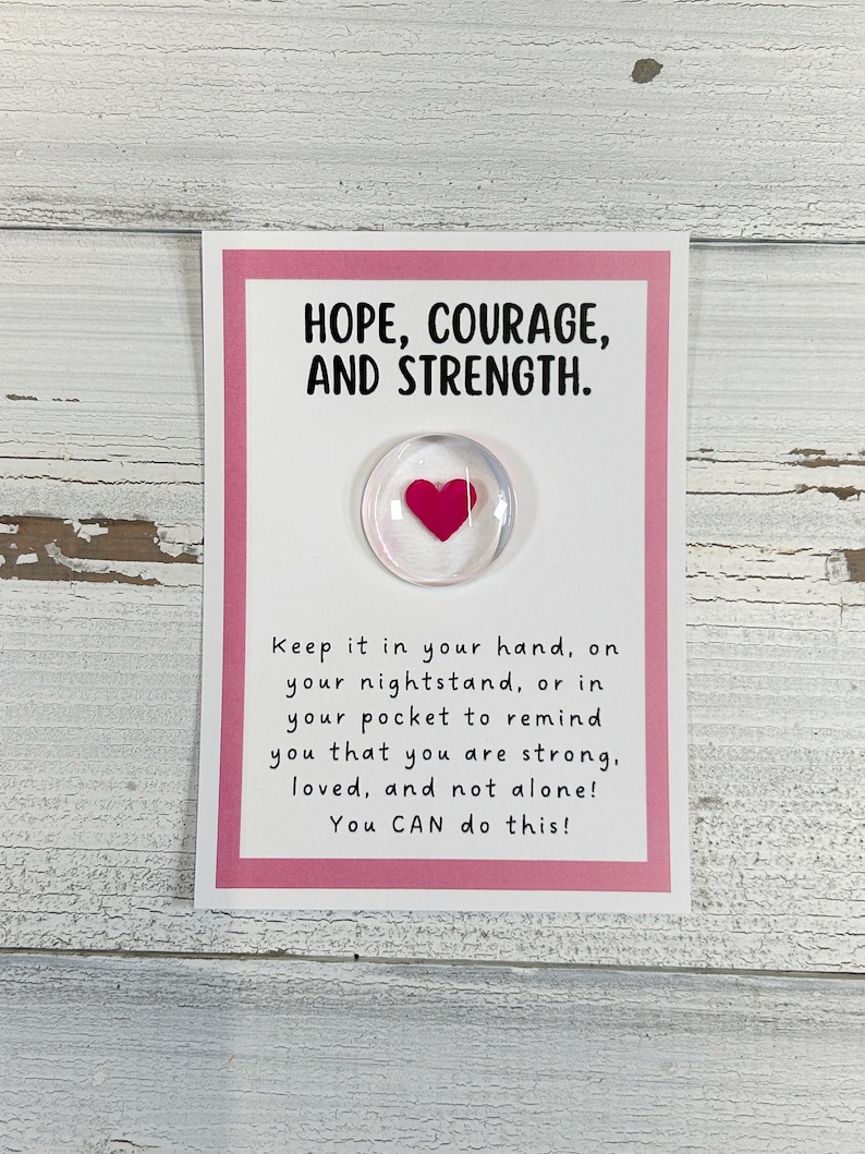 May include: A pink card with the words "HOPE, COURAGE, AND STRENGTH." printed on it. A small glass heart with a pink heart inside is attached to the card. The card reads "Keep it in your hand, on your nightstand, or in your pocket to remind you that you are strong, loved, and not alone! You CAN do this!"
