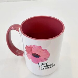 May include: White ceramic mug with a pink flower graphic and the words "Hope Courage Strength" in black lettering. The mug has a pink handle and a pink interior.