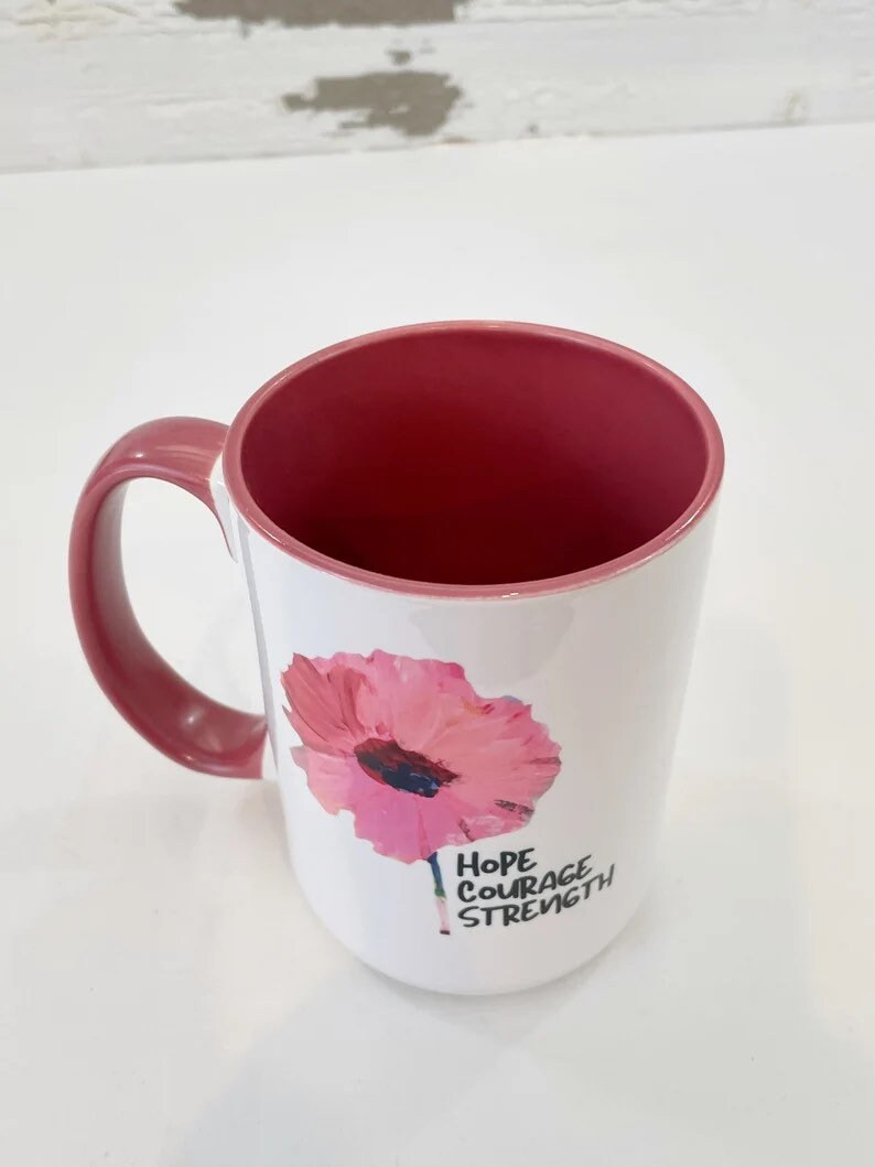 May include: White ceramic mug with a pink flower graphic and the words "Hope Courage Strength" in black lettering. The mug has a pink handle and a pink interior.