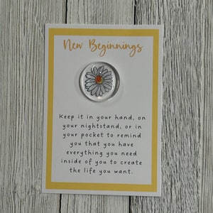 May include: A white card with yellow trim and the text "New Beginnings" printed on it. A small glass circle with a white daisy design is attached to the card. The text below the daisy reads "Keep it in your hand, on your nightstand, or in your pocket to remind you that you have everything you need inside of you to create the life you want."