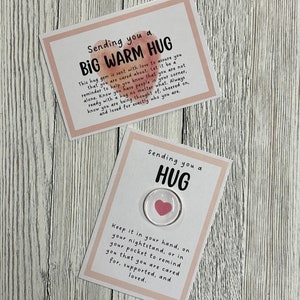 Puede incluir: Dos tarjetas con fondos de acuarela rosa. La tarjeta superior dice "Sending you a BIG WARM HUG" y la tarjeta inferior dice "Sending you a HUG". La tarjeta inferior tiene un peque&ntilde;o amuleto de coraz&oacute;n de vidrio con un coraz&oacute;n rosa en su interior.